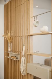 Parete divisoria modulare WoodUpp in rovere a listelli verticali con ripiani sospesi in legno chiaro, ganci metallici e asta porta-tazze, e un contenitore/cassetto metallico beige montato a incastro; design minimal, finitura naturale, adatta ad appendere borse e piccoli oggetti d'arredo.