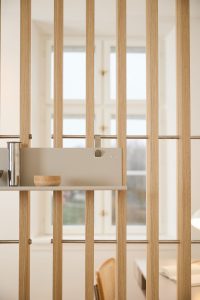 Separatore modulare WoodUpp con listelli verticali in rovere chiaro e barre orizzontali in metallo; mensola/pannello grigio sospeso con accessori WoodUpp visibili: una ciotola tonda in legno e un contenitore cilindrico in acciaio, scorcio luminoso di una stanza retrostante dal design minimale.