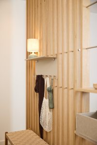 Parete dogata in rovere WoodUpp con pannelli verticali; davanti, appendiabiti a parete WoodUpp color sabbia con mensola superiore e cinque ganci, che reggono sciarpa, borsa a rete e ombrello pieghevole; sulla mensola una lampada con base in ceramica e paralume chiaro.