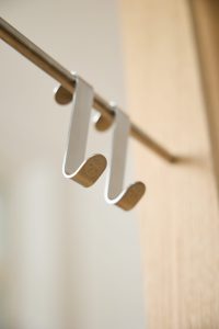 Due ganci appendiabiti WoodUpp in metallo satinato, dal profilo sottile a forma di S, sospesi su una barra metallica; estremità arrotondate con logo inciso a cerchio, montati davanti a un pannello in legno chiaro. Foto ravvicinata, atmosfera minimal e luci morbide.