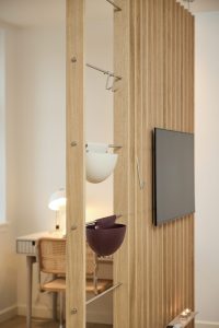 Divisorio verticale in listelli di rovere chiaro con TV montata; due contenitori sospesi WoodUpp (una ciotola bianca e una bordeaux) appesi a barre metalliche e alcuni ganci in acciaio. Sullo sfondo scrivania con lampada e sedia in paglia, dal design moderno e funzionale, illuminata da luce calda.