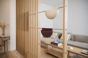 Paravento a lamelle in rovere WoodUpp con aste metalliche su cui sono appesi due vasi WoodUpp a mezzaluna: uno beige opaco e uno bordeaux scuro, dal design sospeso e minimal, che fungono da contenitori decorativi davanti a un soggiorno moderno con divano e tavolino.