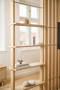 Mobile divisorio WoodUpp in rovere chiaro: struttura a listelli verticali con ripiani a giorno fissati da tiranti metallici, che funge da libreria e separé. Sui ripiani si vedono un piccolo cesto intrecciato, vaso cilindrico in metallo, ciotola d’acciaio, libri e gomitoli di lana.