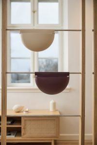 Due vasi WoodUpp a mezzaluna, uno beige chiaro e uno bordeaux scuro, appesi a barre metalliche inserite in una struttura verticale in legno. Contenitori semicircolari dal design minimale e superficie opaca, ideali come portapiante o portaoggetti sospesi. Con fissaggi metallici laterali e linee pulite, perfetti per soggiorno o ingresso.