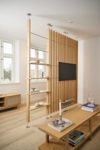 Separé in rovere firmato WoodUpp con listelli verticali e aste a tensione in acciaio inox, dotato di mensole a vista e supporto TV integrato; completa la scena un tavolino basso e un mobile contenitore coordinato in legno chiaro, atmosfera minimal e luminosa.