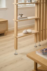 Separé-scaffale WoodUpp in rovere chiaro con listelli verticali e piedi metallici regolabili, due ripiani con gomitoli di lana, libri e una ciotolina metallica; in primo piano un tavolino basso WoodUpp e, sullo sfondo, un mobile contenitore basso WoodUpp con anta in paglia di Vienna.