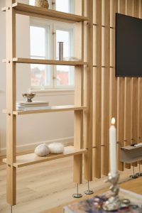 Separatore WoodUpp in rovere chiaro: parete divisoria a lamelle con mensole aperte integrate, poggiata su piedini metallici regolabili. Sulle mensole libri, ciotola argentata e gomitoli di lana; sul lato schermo TV. In primo piano candela accesa e finestra sullo sfondo.
