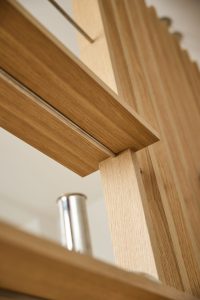 Primo piano di un prodotto WoodUpp: separè modulare in rovere chiaro con doghe verticali regolari e mensole orizzontali. Si vedono traverse metalliche sottili e un supporto cilindrico in acciaio satinato; dettagli sulle giunzioni, venatura naturale del legno e finitura liscia.