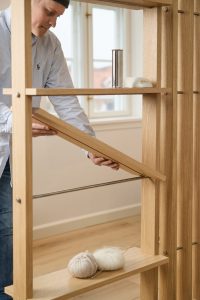 Persona che inserisce un pannello ripiano nella libreria modulare in rovere WoodUpp: struttura a listelli verticali, ripiani regolabili, barrette metalliche e perni in acciaio. In primo piano gomitoli di lana e ferri da maglia appoggiati su un ripiano, luce naturale da finestra.