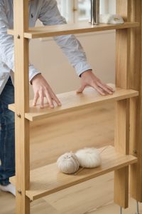 Scaffale modulare in rovere WoodUpp con mensole regolabili, viti metalliche e finitura naturale; mani di una persona che sistemano una mensola. Sulla mensola inferiore due gomitoli di lana beige e bianco con un ferro da maglia, dettaglio della venatura e della struttura a colonna.