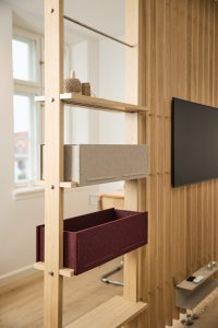 Separé modulare WoodUpp in legno chiaro di rovere con ripiani dal design minimal; due contenitori portaoggetti WoodUpp in feltro compatto (grigio chiaro e bordeaux) inseriti sui ripiani, bobine di spago sulla mensola superiore, finestra sullo sfondo, parete a doghe e televisore parzialmente visibile.