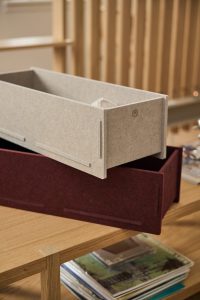 Due organizer WoodUpp in felt: uno beige chiaro posato sopra uno bordeaux scuro, scatole rettangolari modulari con incastri laterali e logo discreto sul pannello anteriore. Appoggiate su un tavolo in rovere, mostrano trama morbida e linee minimal, con riviste impilate sotto.