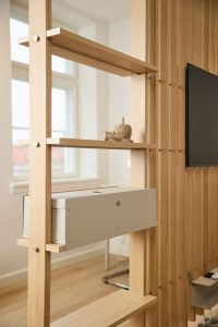 Struttura divisoria modulare WoodUpp in rovere chiaro con ripiani a gradini, scatola portacavi metallica WoodUpp montata su uno scaffale e piccoli oggetti decorativi (spole di corda). Sullo sfondo parete dogata in legno e televisore; linee pulite e minimaliste, viti metalliche a vista e una finestra luminosa.