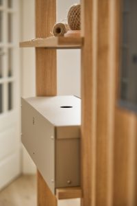 Scaffale modulare WoodUpp in rovere chiaro con montanti verticali e ripiani aperti; contenitore metallico WoodUpp beige fissato al telaio, con foro circolare sul coperchio, piccole viti a vista e foro di aerazione laterale. Sul ripiano superiore spago arrotolato come elemento decorativo.