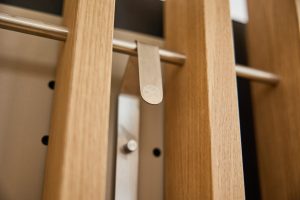 Primo piano di un elemento WoodUpp: listelli verticali in legno chiaro (rovere), una barra metallica orizzontale con un appendino/clip in acciaio satinato inciso con il logo WoodUpp, e una staffa metallica di supporto visibile sullo sfondo, dettagli puliti e finitura naturale che evidenziano le venature del legno.