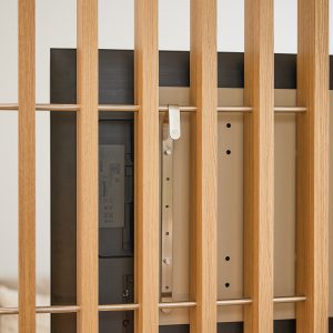 Dettaglio WoodUpp: parete divisoria a listelli verticali in rovere chiaro, con barre orizzontali metalliche, che nasconde il retro di un televisore; si vede la staffa di montaggio WoodUpp in metallo avvitata al pannello, con gancio regolabile per il fissaggio del TV.