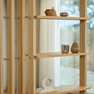 Scaffale modulare WoodUpp in rovere chiaro: montanti verticali a listelli, ripiani aperti e barre in acciaio inox con viti metalliche. Su due ripiani si notano ciotole e contenitori in legno, una tazza metallica e una scultura in marmo a forma di anello.