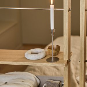 Dettaglio di una mensola divisoria WoodUpp in rovere chiaro con montanti metallici; sopra si vedono un sottile portacandela minimalista in acciaio inox con candela bianca accesa, una ciotola tonda in legno e un oggetto decorativo circolare in marmo. In basso pieghe di tessuto neutro.