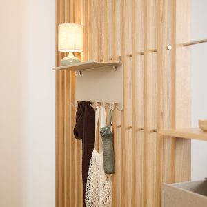Immagine mostra un pannello WoodUpp a doghe in rovere chiaro affiancato da un accessorio WoodUpp: appendiabiti da parete in metallo color sabbia con ripiano superiore e cinque ganci, su cui sono appesi borse, ombrello e sciarpa; una lampada è appoggiata sul ripiano.