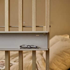 Separatore modulare WoodUpp in listelli di rovere chiaro, dal design minimal e raffinato, con asta metallica e mensolina portaoggetti WoodUpp grigia fissata ai montanti; sulla mensola occhiali da vista e piccolo taccuino, sullo sfondo letto imbottito e atmosfera calda della camera da letto.
