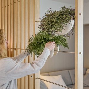 Donna sistema piante in un divisorio WoodUpp: pannello di doghe in legno chiaro WoodUpp con aste in acciaio e vasi sospesi bianchi semi-sferici WoodUpp, contenenti piante verdi rigogliose. Arredo modulare e funzionale che separa ambienti e ospita fioriere sospese dal design contemporaneo elegante.