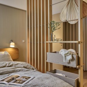 Interno camera: elegante separé WoodUpp in listelli di legno chiaro con scaffali aperti, barre appendiabiti in metallo e due cassetti portaroba in tessuto grigio WoodUpp; sullo scaffale vaso con rami, piccolo piatto decorativo, mentre in primo piano letto sfatto con coperta e libro aperto.