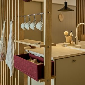 Immagine mostra una cucina con struttura divisoria WoodUpp in rovere: mensole sottili, una barra porta-tazze con ganci che reggono cinque tazze bianche, un ampio cassetto organizer in feltro bordeaux inserito nella struttura e ganci laterali con strofinacci, sopra ripiano e lavello.