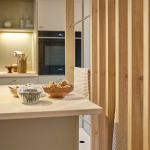 Interno cucina con isola chiara e uno scaffale divisorio WoodUpp: un pannello a listelli verticali in legno (rovere) con barre portasalviette in metallo integrate. Sul piano ciotole in legno con tuberi e una zuccheriera di vetro, asciughino rigato appeso alla barra.