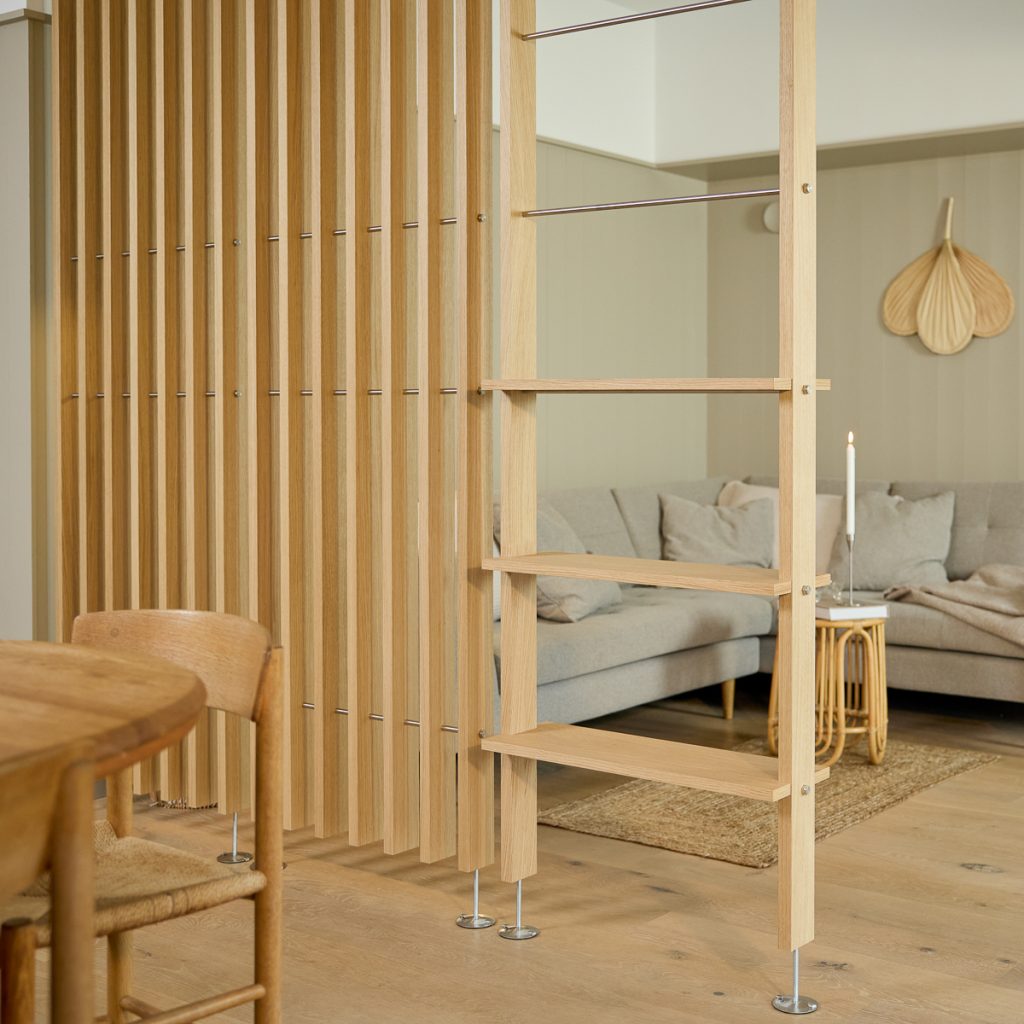 Parete divisoria WoodUpp composta da listelli verticali in rovere chiaro e una struttura libreria a scala WoodUpp con ripiani aperti, tiranti metallici e piedini regolabili, che separano con stile la zona pranzo dal soggiorno con divano grigio e tavolino in rattan.