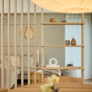 Mobile divisorio e mensola WoodUpp in rovere chiaro con listelli verticali e ripiani aperti; sulle mensole oggetti decorativi: coppetta in legno, contenitore a forma di pera, tazza metallica e scultura circolare in marmo; candela alta accesa e salotto dai toni neutri sullo sfondo.