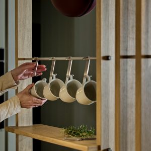 Mobile pensile WoodUpp in legno chiaro con ripiano e struttura a montanti, dotato di barra metallica e ganci WoodUpp che reggono quattro tazze di ceramica bianche; mani che appendono una tazza e rametto di erbe appoggiato sul ripiano, atmosfera cucina minimal.