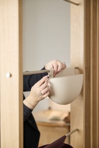 Mani agganciano un vaso pensile beige di WoodUpp, contenitore in plastica opaca a semisfera fissato a una barra metallica; sullo sfondo si vede la struttura appendiabiti in legno chiaro WoodUpp con barra per appendere capi e accessori, dal design minimalista.