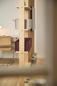 Pannello divisorio WoodUpp in legno di rovere con ripiani a incastro e due contenitori in feltro WoodUpp (grigio chiaro e bordeaux), supporto TV sottile, piccolo ripiano laterale con telecomando e candele. Vista parziale di scrivania e sedia in paglia sullo sfondo.