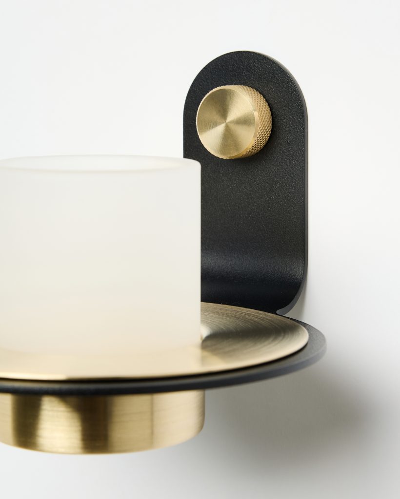 Applique da parete WoodUpp: porta-tealight moderno con base circolare in ottone spazzolato, supporto nero curvo in metallo e cilindro in vetro satinato per la candela, più una manopola zigrinata in ottone dorato come dettaglio decorativo. Include piatto sottostante in ottone, ritratto in primo piano su sfondo bianco.
