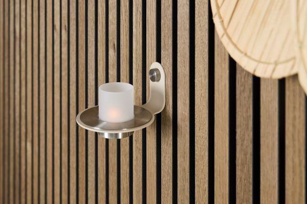 Parete WoodUpp con pannelli a doghe verticali in rovere chiaro e fessure nere; in primo piano un portacandela da parete in metallo satinato con bicchiere opalino e luce calda. In alto a destra si intravede una lampada rotonda in fibra naturale.